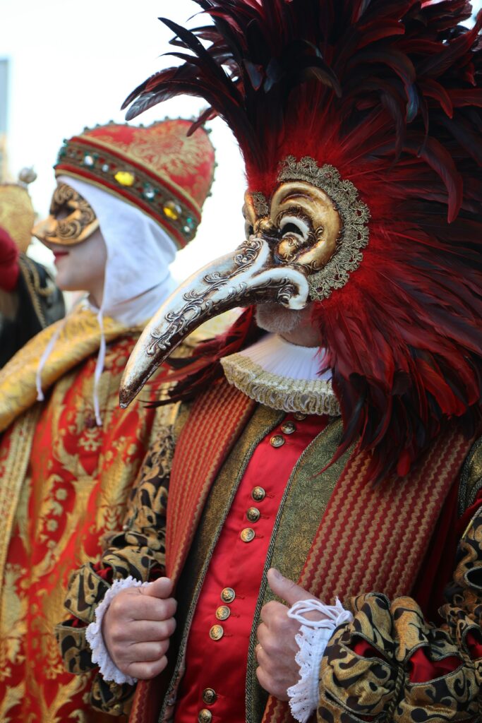 Dos personas con trajes coloridos y ostentosos; usando máscaras del carnaval de Venecia. Predomina el rojo y dorado en la vestimenta.