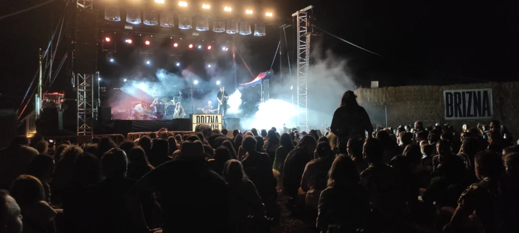Concierto nocturno al aire libre. Una banda toca en un escenario con luces de colores, humo y una estructura de iluminación. El público, de pie y sentado en el suelo, observa la actuación. A la derecha hay un muro de pacas de paja con un cartel que dice «BRIZNA».